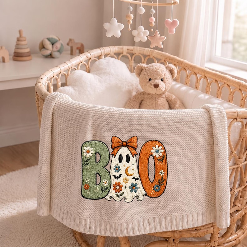 May include: A cream-colored knit baby blanket featuring an embroidered design of the word BOO. The letter B is sage green with floral accents, the letter O is orange with floral accents, and a ghost with a bow and stars replaces the middle O.