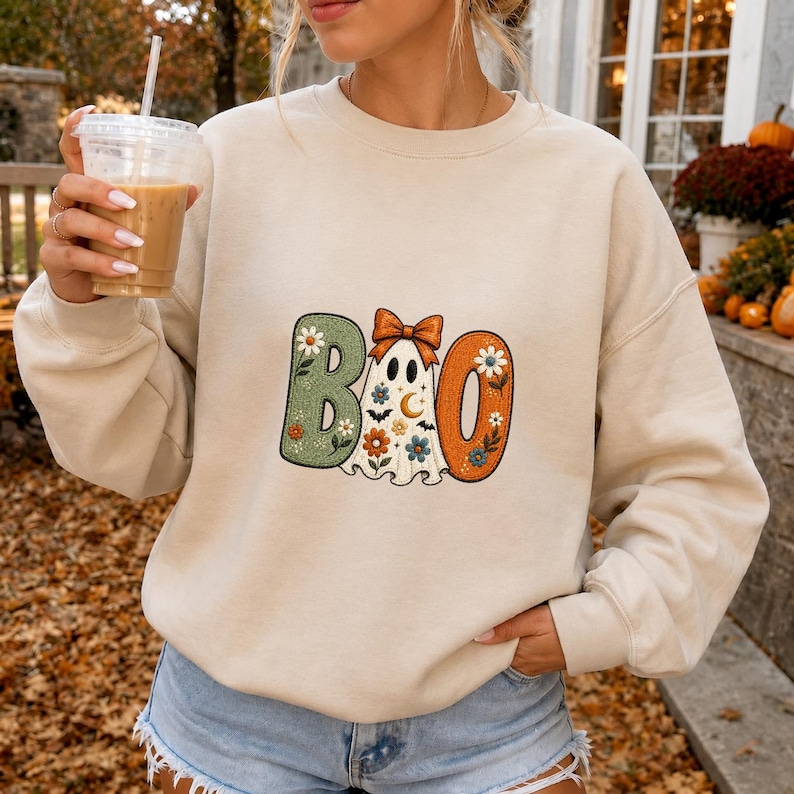 May include: A beige crewneck sweatshirt featuring a graphic print that spells BOO. The letter B is sage green with floral patterns, the letter O is a white ghost wearing an orange bow with stars and bats, and the final O is orange with flowers.