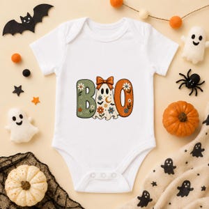 May include: A white short-sleeved baby bodysuit featuring a graphic print of the word BOO. The letter B is sage green with floral patterns, the letter O is orange with floral patterns, and the ghost in the middle wears an orange bow.