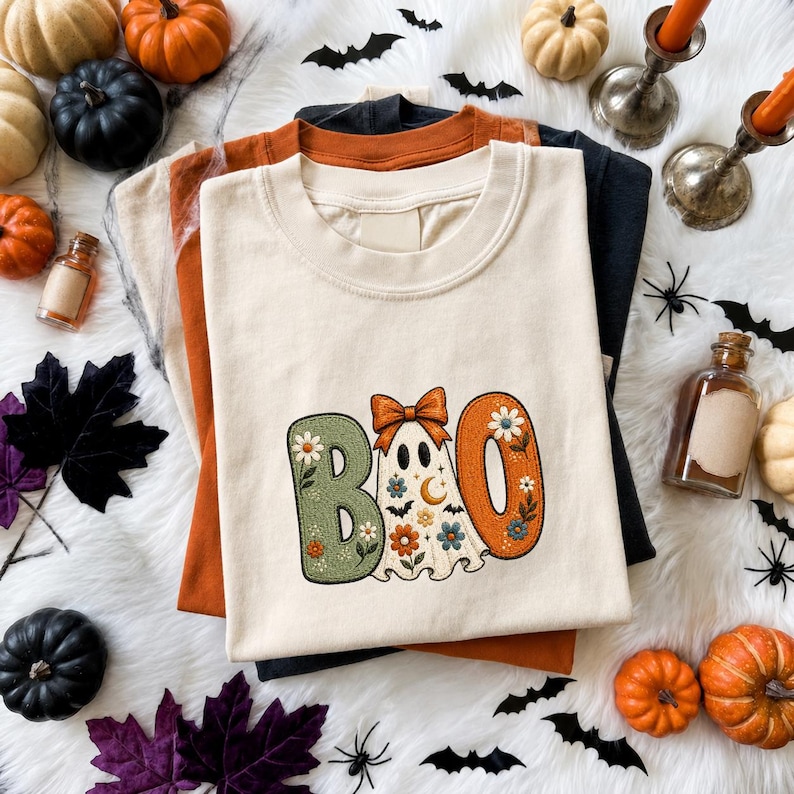 May include: A cream-colored t-shirt featuring a graphic of the word BOO, where the letter O is replaced by a ghost wearing an orange bow. The letters are decorated with floral patterns in green and orange. The shirt is layered over orange and black shirts.