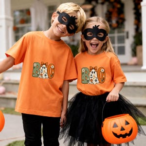 May include: Two orange t-shirts featuring a graphic of a ghost with a bow and the text BOO, decorated with flowers and stars. The shirts are worn with black masks, and one is paired with a black tulle skirt and an orange pumpkin bucket.