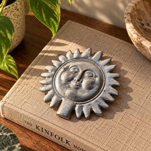 May include: A circular silver-toned metal sun face ornament featuring a stylised human face with closed eyes and prominent cheeks, surrounded by radiating sun rays. The object has a small rectangular base at the bottom and a textured metallic finish.