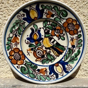 May include: A ceramic plate with a hand-painted design featuring a bird and floral motifs. The plate has a white base with a blue rim and is decorated with orange, blue, green, and yellow details. The bird is perched amidst the flowers.