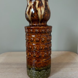 May include: A tall ceramic vase with a textured surface and a unique design. The vase features a brown and tan upper section, a cylindrical middle section with circular accents, and a textured green base. The vase is approximately 30 cm tall.