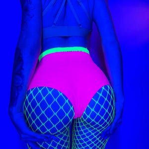 Könnte beinhalten: Hoch taillierter neonpinker Slip mit einem leuchtend lindgrünen Bund, kombiniert mit neon-lindgrünen Netzstrümpfen. Die Kleidungsstücke werden unter ultraviolettem Licht beleuchtet, was einen hellen, leuchtenden Effekt vor einem dunkelblauen Hintergrund erzeugt.