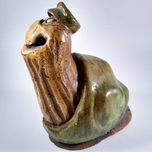 May include: A ceramic sculpture of a stylized creature with a green and brown glaze. The creature has a large head with a pointed beak and a small, round eye. It is wearing a small, green hat.