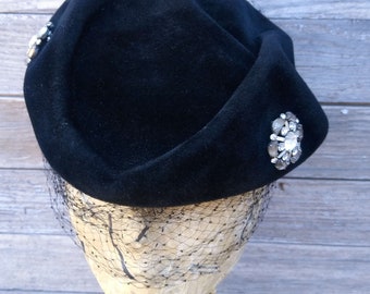 May include: A black velvet hat with a wide brim and a net veil. The hat is decorated with two large, clear rhinestone brooches.