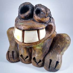 May include: A ceramic frog figurine with a wide, toothy grin. The frog is brown and green with white teeth and black eyes.