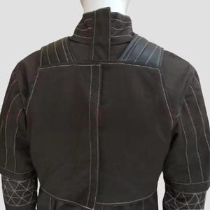 K&ouml;nnte beinhalten: Schwarze Jacke mit wei&szlig;en N&auml;hten. Die Jacke hat einen hohen Kragen, gepolsterte Schultern und geometrische Muster an den &Auml;rmeln. Die R&uuml;ckansicht zeigt die N&auml;hte und Designdetails des Kleidungsst&uuml;cks.