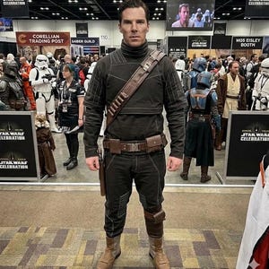 K&ouml;nnte beinhalten: Eine Person in einem schwarzen Kost&uuml;m mit braunen Akzenten, darunter G&uuml;rtel, Holster und Stiefel, steht vor einem Star Wars Celebration Banner. Das Kost&uuml;m hat einen hohen Kragen und gepolsterte Details. Andere Star Wars Charaktere und Teilnehmer sind zu sehen.