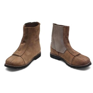 K&ouml;nnte beinhalten: Zwei braune Stiefeletten aus Wildleder mit Rei&szlig;verschluss und schwarzer Sohle. Ein Stiefel hat einen hellbraunen Akzent am oberen Schaft. Die Stiefel werden vor wei&szlig;em Hintergrund gezeigt.