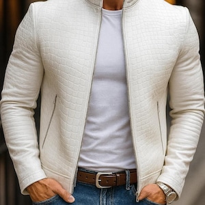 Op de afbeelding: Een witte, getextureerde leren jas met ritssluiting en opstaande kraag. De jas wordt gecombineerd met een wit t-shirt, een blauwe jeans en een bruine leren riem. Een zilveren horloge is zichtbaar om de pols.