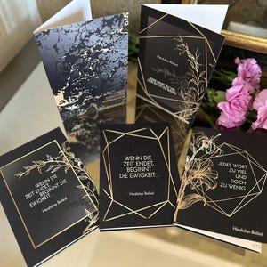 May include: A collection of black greeting cards with gold geometric designs and botanical illustrations. The cards feature text in German, with phrases like "Wenn die Zeit endet, beginnt die Ewigkeit..." and "Jedes Wort zu viel und doch zu wenig."