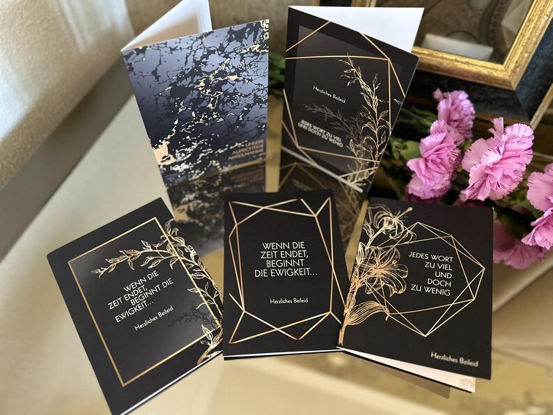 May include: A collection of black greeting cards with gold geometric designs and botanical illustrations. The cards feature text in German, with phrases like "Wenn die Zeit endet, beginnt die Ewigkeit..." and "Jedes Wort zu viel und doch zu wenig."