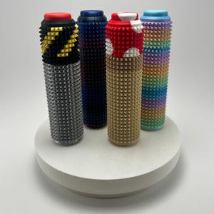 May include: A collection of four cylindrical objects with textured surfaces and colorful tops. The objects are arranged on a white circular platform. Colors include black, blue, red, white, tan, and a rainbow gradient. The objects have a studded pattern.