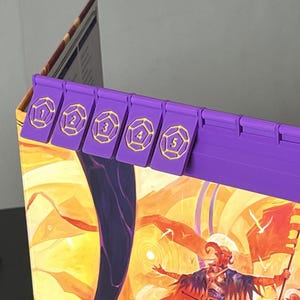 May include: Purple game board dividers with gold numbers 1 to 5. The dividers are attached to the edge of a game board with colourful artwork. The game board features a desert landscape with figures.