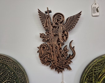 May include: A wooden carving of Saint Michael the Archangel, depicted with wings, a cross, and a shield, standing over a dragon. The carving is a warm brown color and is mounted on a white wall.
