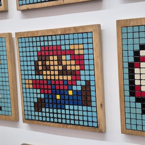 May include: Pixelated art pieces featuring iconic video game characters, including Mario and Luigi, in a mosaic style. Each piece is framed in natural wood, creating a retro aesthetic. The artwork is mounted on a white wall.