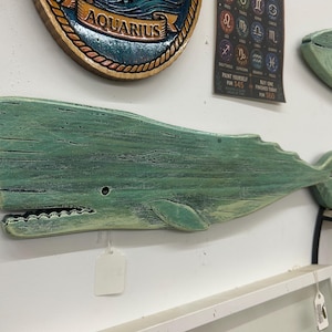 May include: A wall-mounted wooden whale cutout featuring a distressed, weathered teal and green paint finish. The whale has a visible eye, a carved mouth with teeth, a side fin, and a tail. A small white price tag hangs from the lower jaw area.