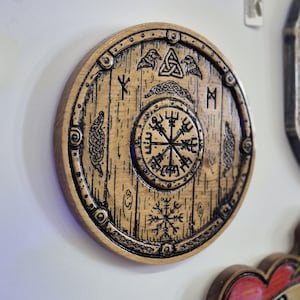 Op de afbeelding: Een ronde, houten wanddecoratie met een gebeeldhouwd Viking schildontwerp. Het schild is bruin met zwarte details, met runen, een kompassymbool en andere Noorse motieven. Het schild heeft een diameter van ongeveer 25 cm.