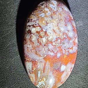May include: Oval-shaped gemstone with a polished surface. The stone features a mix of colors, including white, orange, and red, with intricate patterns. The gemstone is set against a dark background, highlighting its natural beauty.