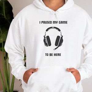 May include: White hoodie featuring a black headphone graphic and the text "I PAUSED MY GAME TO BE HERE". The hoodie has a drawstring hood and a front pocket. The person is wearing the hoodie.