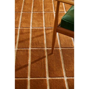 May include: A brown rectangular rug with a grid pattern of brown blocks separated by off-white lines. A wooden chair leg and a green cushion are visible in the upper right corner. The rug has a soft, textured appearance.