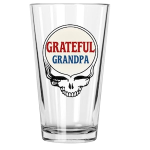 May include: Clear pint glass with a black and white skull design. The design includes a cream-colored circle with the words "Grateful Grandpa" in red and blue lettering. The glass is empty and sits against a white background.
