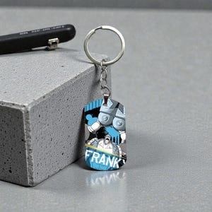 Puede incluir: Un llavero rectangular con un llavero y una cadena plateados. El llavero presenta una ilustración colorida de un personaje con el nombre "FRANK" en azul y blanco. El fondo tiene detalles en azul y gris.