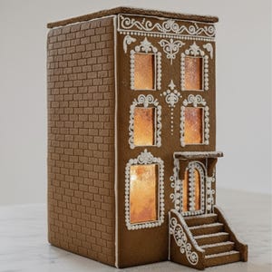 Può includere: Una casa di pan di zenzero con facciata in mattoni e glassa bianca decorativa. La casa ha quattro finestre illuminate e una porta d'ingresso con gradini. Il tetto è piatto e decorato con glassa bianca. Una decorazione festiva.