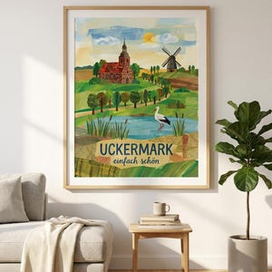 May include: A framed art print featuring a colorful landscape with a church, windmill, and lake. The artwork has a rustic style with the text "UCKERMARK einfach schön" in a navy blue banner. The print is displayed in a living room setting.