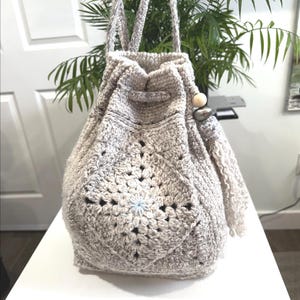May include: A handmade, light gray crochet drawstring bag with a geometric pattern on the front. The bag has a long strap and a decorative tassel with wooden beads. The bag is sitting on a white surface.