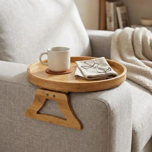 Puede incluir: Una bandeja redonda de bambú para el brazo del sofá con soporte incorporado. Una taza blanca está sobre un posavasos y unas gafas descansan sobre un paño doblado. La bandeja está sujeta al brazo de un sofá gris.