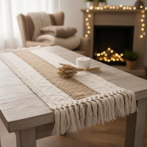 Peut inclure: Chemin de table en macramé crème et beige avec des franges. Le chemin de table présente une bande centrale beige bordée de sections crème. Un petit bouquet de blé et une bougie blanche sont sur la table en bois clair.