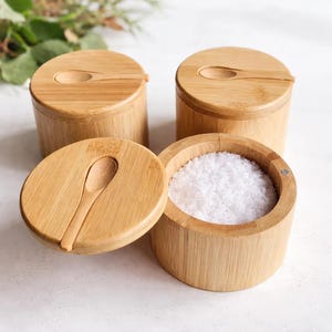 以下が含まれることがあります： 蓋と小さなスプーンが付いた4つの竹製ソルトセラー。1つのセラーが開いており、白い塩が見えます。セラーは円筒形で、天然の木目仕上げです。蓋には小さなスプーン型のくぼみがあります。
