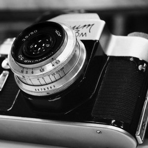 May include: A black and silver 35mm film camera with a lens that has a depth of field scale and a silver lens cap. The camera has a black leather strap.