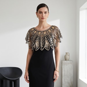 May include: A black sleeveless dress featuring a decorative capelet overlay. The capelet is adorned with a gold and black sequined leaf-like pattern arranged in a radial design around the neckline, extending over the shoulders and upper chest.