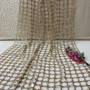 May include: A gold-coloured, open-weave fabric with a grid pattern and small, reflective embellishments. The fabric is draped, with a few sprigs of pink and purple flowers in the corner. The background is a light, patterned wall.
