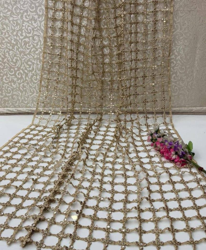 May include: A gold-coloured, open-weave fabric with a grid pattern and small, reflective embellishments. The fabric is draped, with a few sprigs of pink and purple flowers in the corner. The background is a light, patterned wall.
