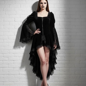 May include: Black velvet dress with a square neckline, long bell sleeves with lace trim, and a high-low hemline. The dress features a front zipper and is styled with black heels. The dress is against a white brick wall.