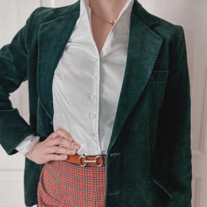 May include: A dark green velvet blazer paired with a white button-down shirt and red plaid pants. A brown belt with a gold buckle cinches the waist. The outfit is a mix of textures and patterns, creating a stylish ensemble.