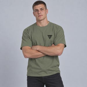 May include: An olive green short-sleeve t-shirt featuring a small black triangular graphic print on the left chest. The shirt has a crew neckline and a standard fit, paired with dark charcoal gray denim jeans.