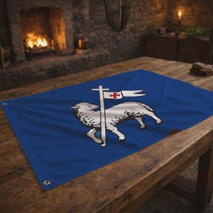 Op de afbeelding: Een blauwe vlag met een wit lam en kruisontwerp. Het lam kijkt naar rechts, met een kruis en banner erboven. De banner heeft een rood kruis. De vlag ligt op een houten tafel.