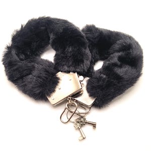 Op de afbeelding: Zwarte kunstbont handboeien met zilverkleurige metalen sluitingen en twee sleutels. De handboeien zijn zacht en pluizig, met een pluche textuur. De sleutels zijn bevestigd aan een kleine metalen ring.