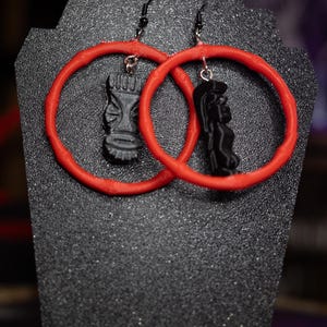 Pode incluir: Um par de brincos pretos em forma de tiki com aros vermelhos. Os brincos são exibidos em uma superfície preta texturizada. As detalhadas figuras tiki estão suspensas dentro dos aros vermelhos vibrantes, criando um contraste marcante.