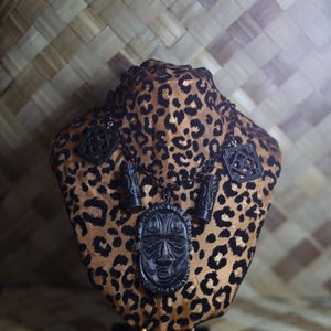 Pode incluir: Um colar preto de estilo tribal com um pingente com um rosto esculpido, exibido em um busto com estampa de leopardo. O colar tem vários amuletos e está sobre um fundo tecido.