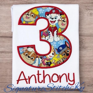 May include: White t-shirt featuring a large red number 3 filled with cartoon images of Paw Patrol characters. The name "Anthony" is embroidered in red below the number. The text "Signature StitchKy" is at the bottom.