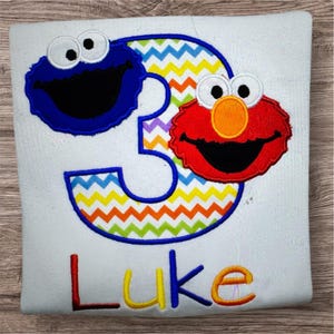 May include: White t-shirt with a colorful number 3 design featuring Cookie Monster and Elmo characters. The number 3 has a zig-zag pattern in multiple colors. The name "Luke" is embroidered in colorful letters.