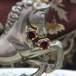 Pode incluir: Brincos de botão em forma de coração com uma pedra preciosa vermelha escura e uma borda dourada com contas. Os brincos são exibidos em uma estátua de cavalo prateada, criando um contraste de texturas e cores.