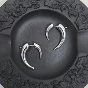 Puede incluir: Un par de pendientes plateados con forma de cuerno. Los pendientes curvados tienen un acabado pulido. Se exhiben en un plato decorativo gris oscuro con un patrón floral en los bordes.